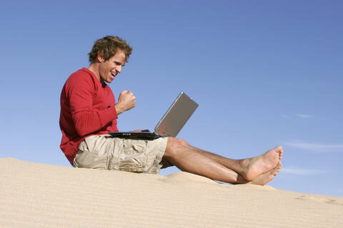 Mann sitzt mit Laptop in der Wüste im Sand und ballt die Faust, lizenzfreies Stockfoto