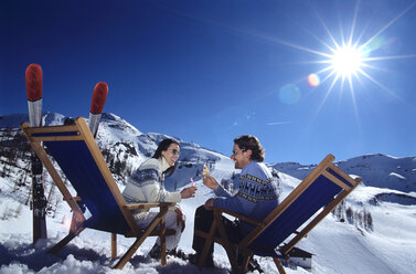 Paar sitzt auf einem Liegestuhl im Schnee und stößt mit Gläsern an, Seitenansicht - HHF00190