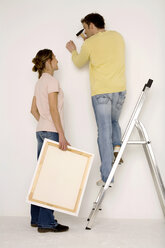 Young couple hanging up painting, man standing on ladder - WESTF00585