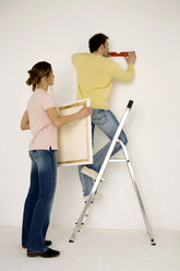 Young couple hanging up painting, man standing on ladder - WESTF00584