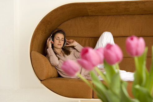 Frau entspannt auf Sofa mit Kopfhörern, rosa Tulpen im Vordergrund - WESTF00549
