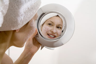 Young woman applying face mask looking into mirror smiling, close-up - MAEF00059