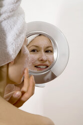 Young woman applying face mask looking into mirror smiling - MAEF00058
