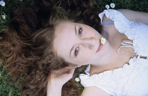 Junge Frau mit Gänseblümchen im Mund im Gras liegend, Nahaufnahme, Porträt - WESTF01166