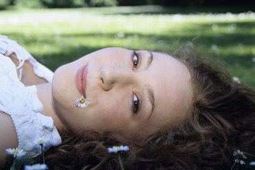 Junge Frau mit Gänseblümchen im Mund im Gras liegend, Nahaufnahme, Porträt - WESTF01165