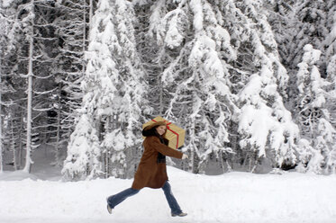Junge Frau trägt Weihnachtsgeschenk auf den Schultern im Schnee, lächelnd - HHF00465