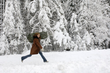 Junge Frau trägt Weihnachtsbaum auf den Schultern im Schnee, lächelnd - HHF00462