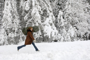 Young woman carrying Christmas tree on shoulders in snow, smiling - HHF00462