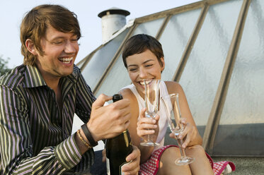Junger Mann öffnet eine Flasche, während eine Frau mit einem Glas sitzt und lächelt - WEST01535