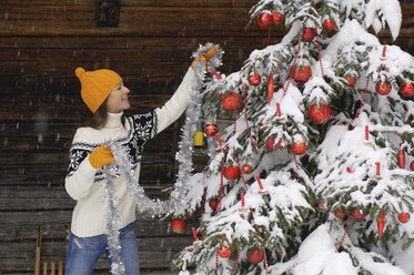 Young woman decorating Christmas tree in snow, smiling - HHF00534