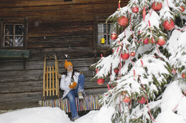 Woman sitting in front of mountain shelter, looking at Christmas tree - HHF00532