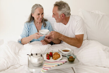 Älteres Paar sitzt mit Frühstück auf dem Bett, lächelnd - WESTF01876
