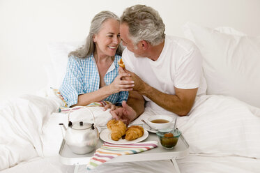 Älteres Paar sitzt mit Frühstück auf dem Bett, lächelnd - WESTF01875
