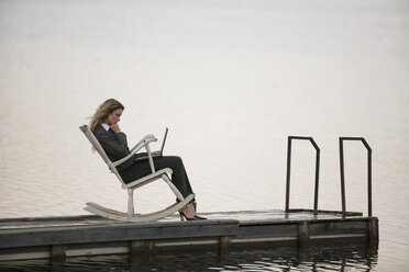 Geschäftsfrau sitzt im Schaukelstuhl auf dem Steg und benutzt einen Laptop, Seitenansicht - WESTF03063