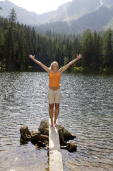 Junge Frau steht auf einer Holzplanke am See und streckt die Arme aus - HHF00790