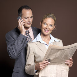 Businessman using mobile phone businesswoman holding newspaper, smiling, portrait - JLF00222