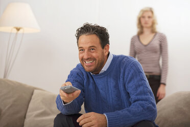 Man sitting on sofa, holding remote control, daughter in background - WESTF03387
