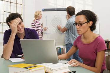 Young couple standing by flipchart, focus on man and woman using laptop in foreground - WESTF03627