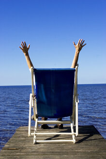 Woman sitting in deck chair on jetty, arms up, rear view - LDF00396