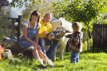 Eltern mit Kind sitzen im Garten und spielen mit Pony - HHF01198