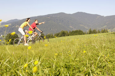 Young couple riding mountain bike in meadow, side view, mountains in background - WESTF04209