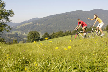 Young couple riding mountain bike in meadow, side view, mountains in background - WESTF04207