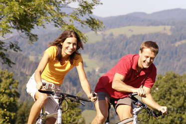 Junges Paar fährt Fahrrad, lächelnd, Berge im Hintergrund - WESTF04206