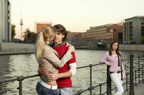 Paar umarmt sich am Fluss, junge Frau steht im Hintergrund, lizenzfreies Stockfoto