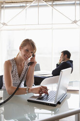 Business woman using laptop while phoning, male colleague in background - WESTF05598