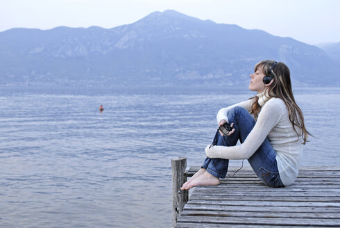 Italien, Gardasee, Junge Frau (20-25) auf Steg sitzend mit persönlicher Stereoanlage - DKF00123