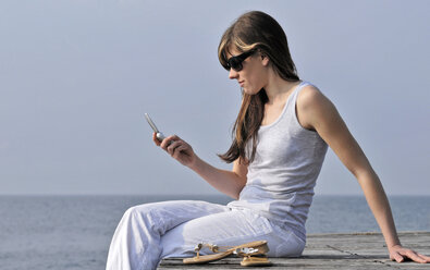 Italy, Lake Garda, Womann 20-25)  sitting on edge of dock with mobile phone, side view - DKF00121