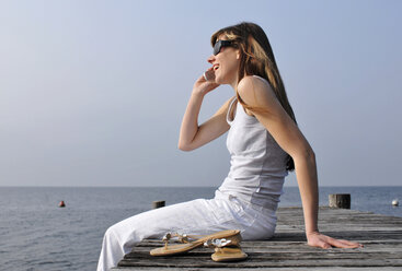 Italy, Lake Garda, Womann 20-25)  sitting on edge of dock with mobile phone, side view - DKF00120