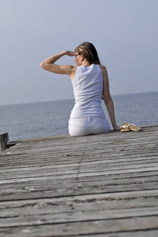 Italy, Lake Garda, Young woman (20-25) sitting on dock, rear view, close-up, lizenzfreies Stockfoto