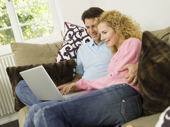 Young couple on sofa using laptop, smiling - WESTF06563