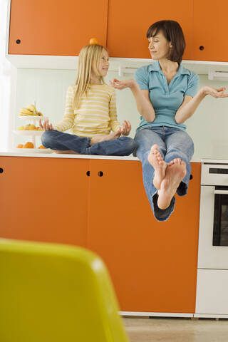 Mutter und Tochter (8-9) in der Küche, Tochter balanciert Orange auf dem Kopf, lizenzfreies Stockfoto
