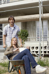 Junges Paar auf einer Baustelle, Frau sitzt in einer Schubkarre - WESTF09158