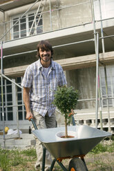 Junger Mann auf einer Baustelle, der eine Schubkarre mit Pflanzen schiebt - WESTF09155