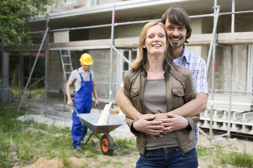Junges Paar auf der Baustelle, das sich umarmt, Bauarbeiter im Hintergrund - WESTF09153