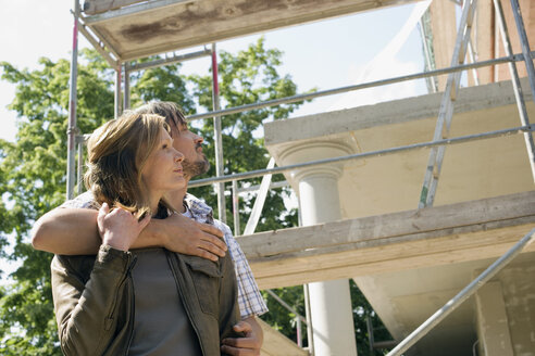 Junges Paar umarmt sich vor einem im Bau befindlichen Haus - WESTF09136