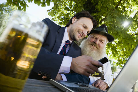 Germany, Bavaria, Upper Bavaria, Senior Bavarian man and young businessman with laptop in beer garden stock photo