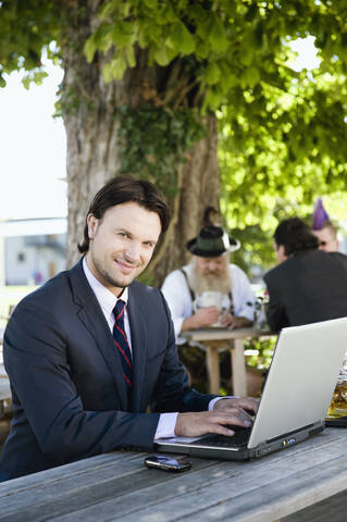 Deutschland, Bayern, Oberbayern, Junger Geschäftsmann im Biergarten mit Laptop, lächelnd, Portrait, lizenzfreies Stockfoto