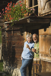 Austria, Karwendel, Senior couple leaning on log cabin, holding mugs - WESTF10469