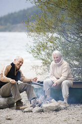 Germany, Bavaria, Senior couple sitting at campfire, grilling fish - WESTF10221