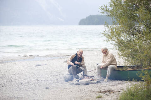 Germany, Bavaria, Senior couple sitting at campfire, grilling fish stock photo
