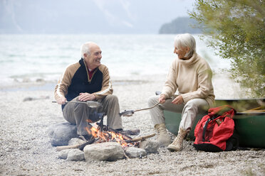 Germany, Bavaria, Senior couple sitting at campfire, grilling fish - WESTF10219