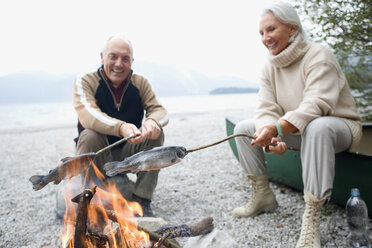 Germany, Bavaria, Senior couple sitting at campfire, grilling fish - WESTF10218