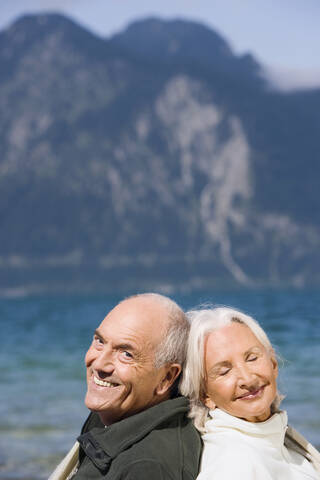 Germany, Bavaria, Senior couple relaxing on lakeshore, sitting back to back, smiling, portrait stock photo