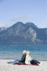 Germany, Bavaria, Senior couple relaxing on lakeshore, sitting back to back - WESTF10141