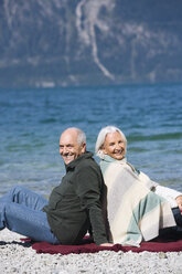 Deutschland, Bayern, Älteres Paar entspannt am Seeufer, Rücken an Rücken sitzend - WESTF10140