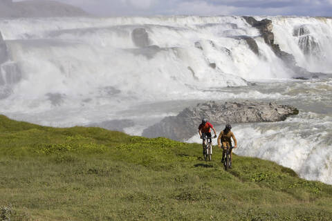 Island, Männer beim Mountainbiking, Wasserfall im Hintergrund, lizenzfreies Stockfoto